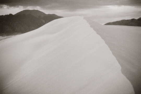 A black-and-white landscape photograph of the ridge of a sand dune against dark mountains and cloudy sky.