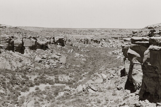 A black-and-white photograph of a scrubby desert landscape framed by high rocks and cliffs.