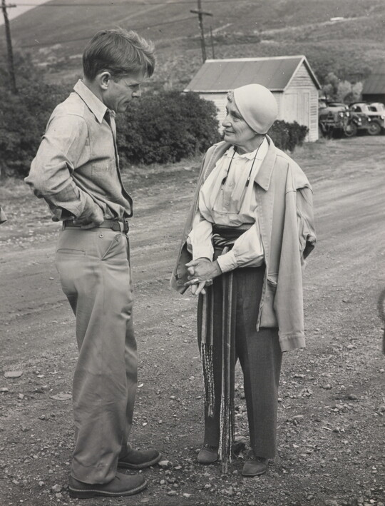A black-and-white photograph of a White man and a White woman talking to each other on a gravel road.