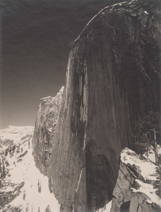 A black-and-white photograph of a steep cliff face and the snow-covered mountains around it.