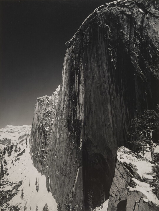 A black-and-white photograph of a steep cliff face and the snow-covered mountains around it.