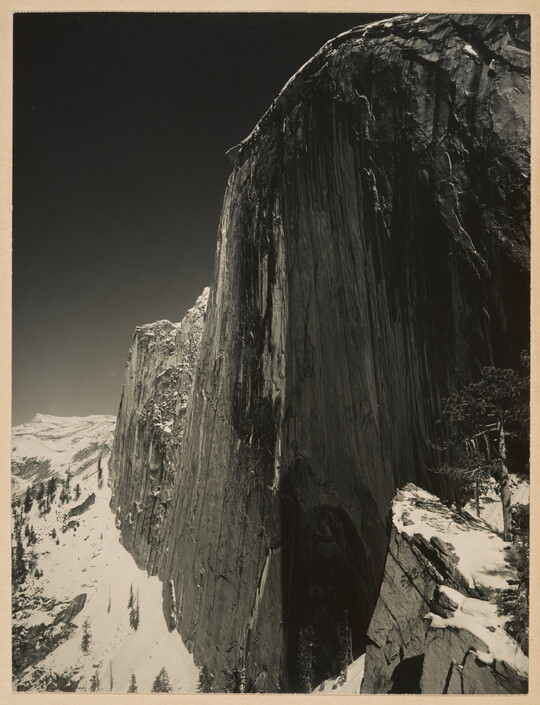 A black-and-white photograph of a steep cliff face and the snow-covered mountains around it.