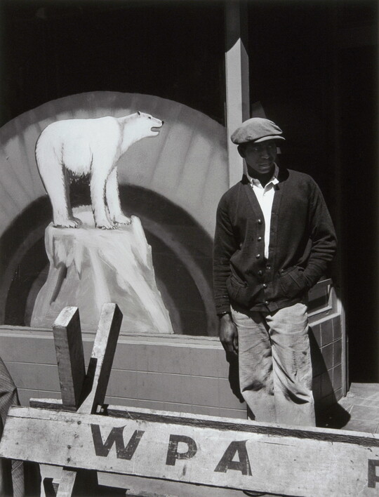 A black-and-white photograph of a Black man in a flat cap and dark cardigan behind a wooden barricade that says "WPA" and in front of a sign with a painting of a polar bear.