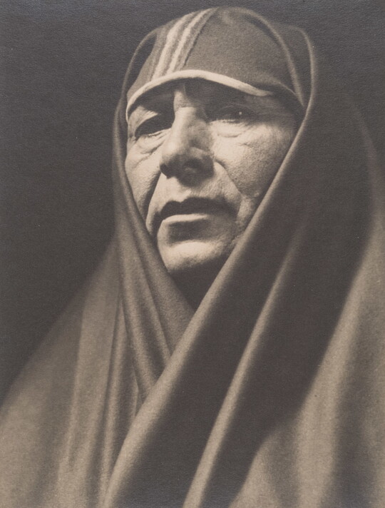 A sepia-toned portrait photograph of an adult Indigenous person with a shawl covering their head and shoulders.