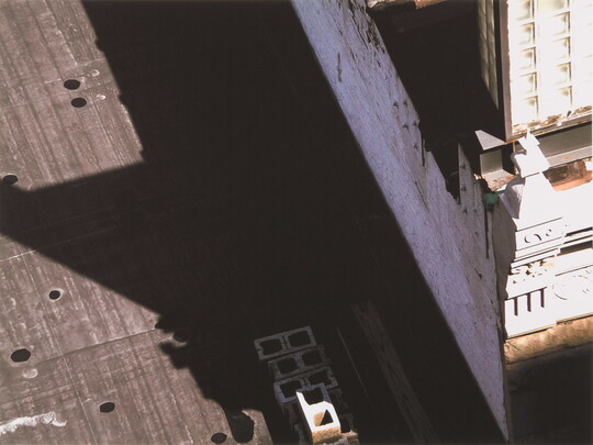 A black-and-white photograph of a building and the shadow it casts across the top of a neighboring building as viewed from the above.
