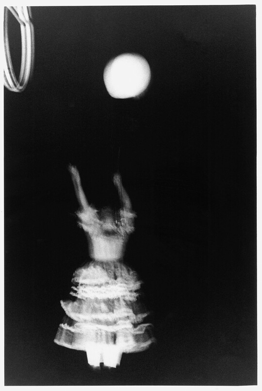 A black-and-white photograph of a person in motion wearing a white dress with a ruffled skirt, arms straight up as if reaching for the circular object above them.