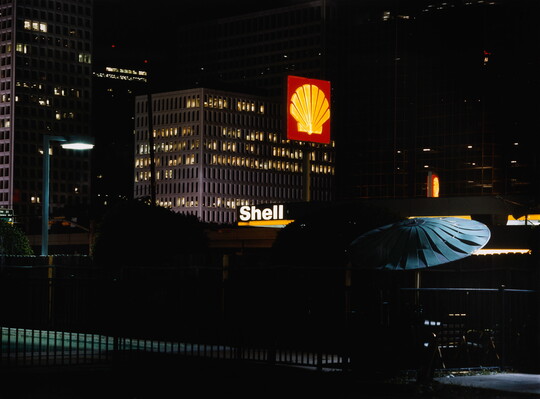 A color photograph of a city at night from a nearby rooftop patio with a Shell gas station sign lit up.