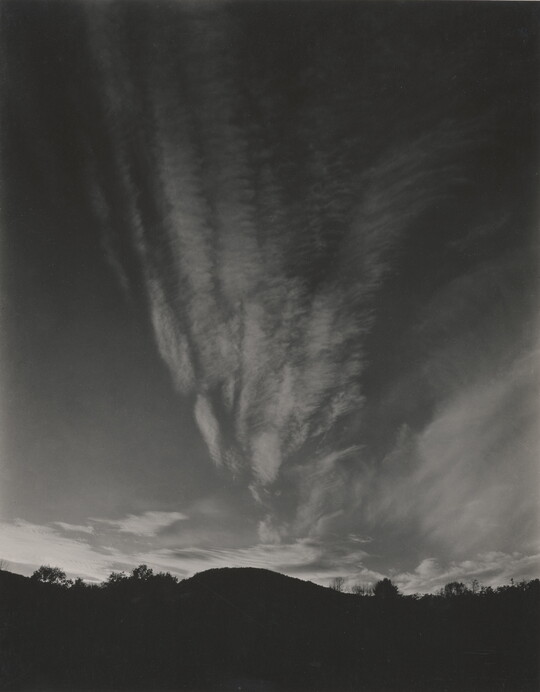 A black-and-white photograph of hills and trees silhouetted against a sky with thin, vertical clouds.