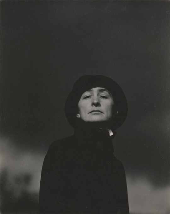 A black-and-white photograph looking up at a young White woman, who is looking down at the camera, wearing a black hat and top.