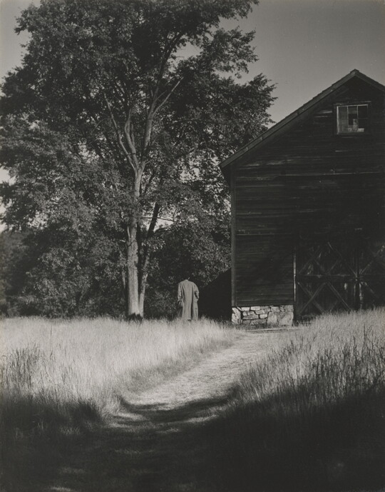 A black-and-white photograph of a person walking on a grassy path that leads to large barn next to a tree.