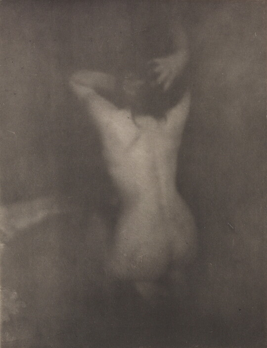 A dark, soft-focus, black-and-white photograph of a nude White woman kneeling with her back to the viewer and her hands in her hair.