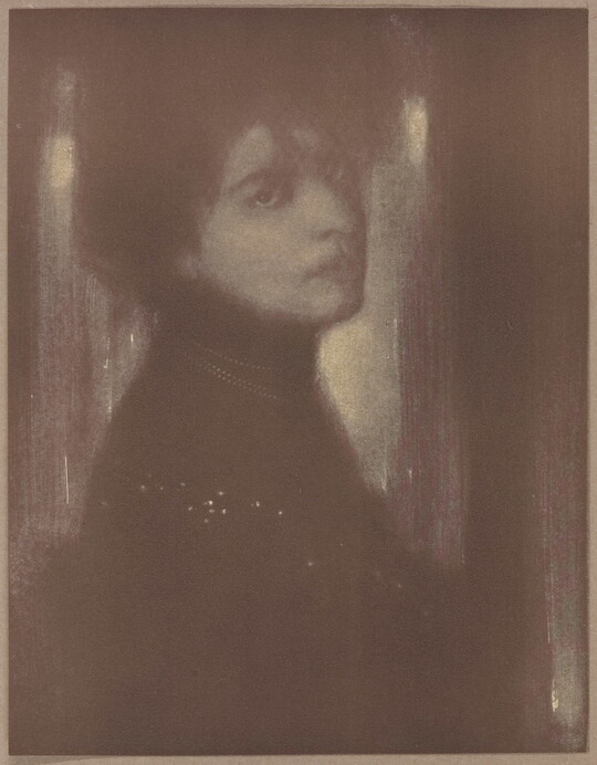 A dark, soft-focus black-and-white portrait photograph of a White woman with her hair up and head turned over her shoulder to face the camera.