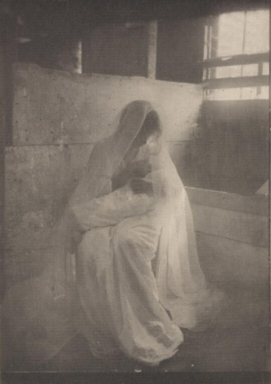 A soft-focus, black-and-white photograph of a seated woman wearing a white dress and veil, holding a baby wrapped in a white blanket.