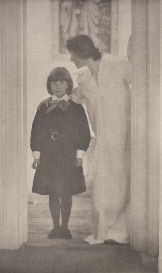 A soft-focus, black-and-white photograph of a White woman in a long, white dress with her arm around the shoulders of a White girl wearing a dark dress standing in a door frame.