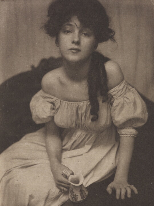A black-and-white portrait photograph of a seated young White woman dressed in white and leaning forward to gaze into the camera.