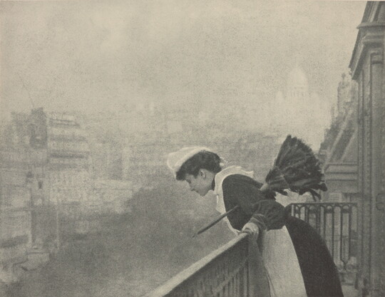 A black-and-white photograph of a White woman dressed as a maid in a white cap, black dress, and white pinafore holding a feather duster under her arm as she peers over a balcony at a smoggy cityscape.