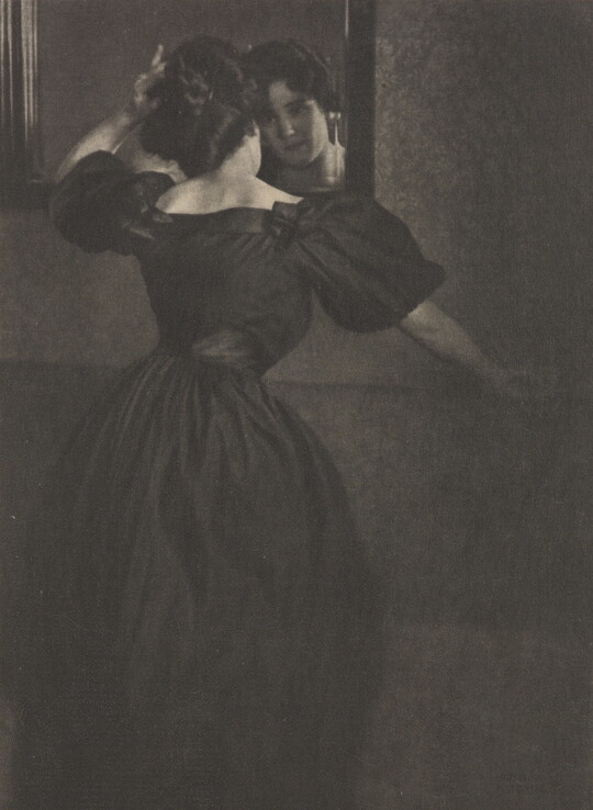 A black-and-white photograph of a young White woman, back to the camera, wearing a long, black dress with puffy sleeves fixing her hair in a mirror.