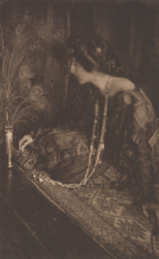 A sepia-toned photograph of a seated White woman wearing a long, patterned dress and a long, beaded necklace leaning toward a vase filled with peacock feathers.