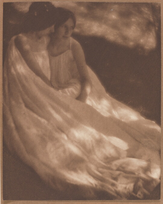 A sepia-toned photograph of two White women, both wearing long flowy dresses, sitting close together on the ground.