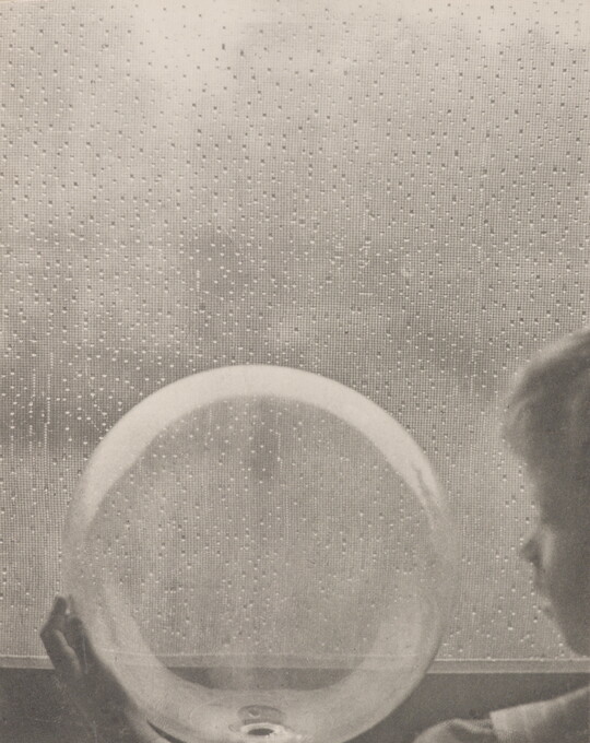 A black-and-white photograph of a White child looking at a clear glass globe in front of a rain-spattered window.
