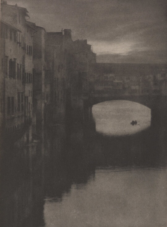 A sepia-toned photograph of a wall of tall buildings lining a river and an enclosed arched bridge stretching over the water.
