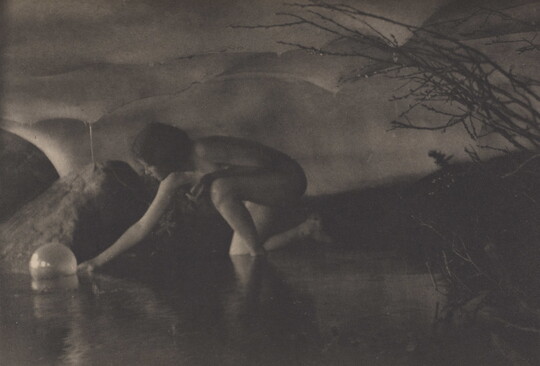 A black-and-white photograph of a nude White women reaching down to pick up a sphere in a shallow body of water.