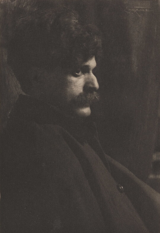 A black-and-white photograph of a White man with lots of dark, curly hair and a mustache sitting in profile.