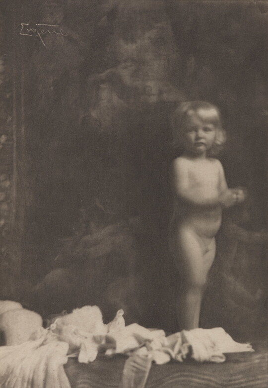 A sepia-toned photograph of a nude White toddler standing next to what looks like a pile of light-colored clothes.