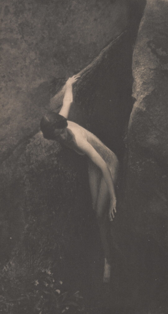 A sepia-toned photograph of a nude White woman emerging from between two large boulders.