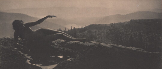 A sepia-toned photograph of a nude woman in shadow lying on a hilltop with cascading hills behind her.