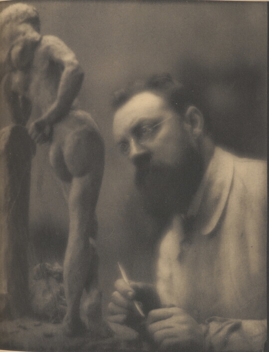 A sepia-toned photograph of a White man with glasses and a beard, holding a sculpting tool in his hand, looking a small sculpture of a nude woman.