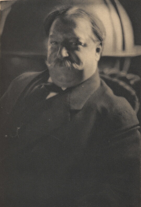 A sepia-toned portrait photograph of a White man with a curled mustache wearing a suit jacket and tie.