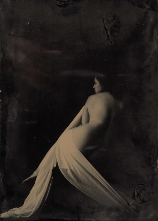 A black-and-white photograph of a seated nude White woman lightly draped in fabric with her back to the camera.