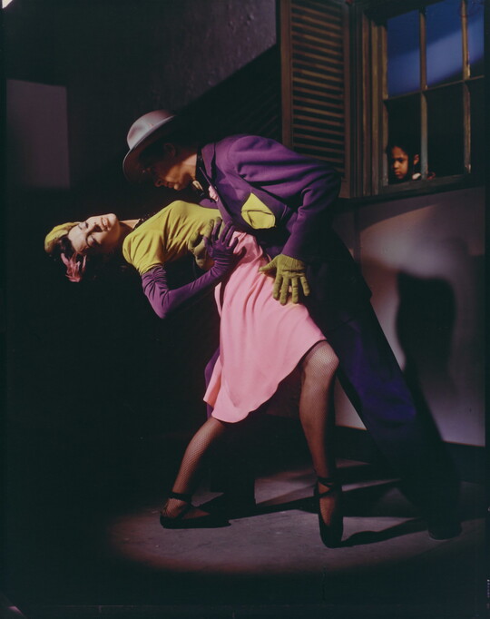 A color photograph of a White man and woman dancing in a dark room in formal attire while a Black child watches through a window.