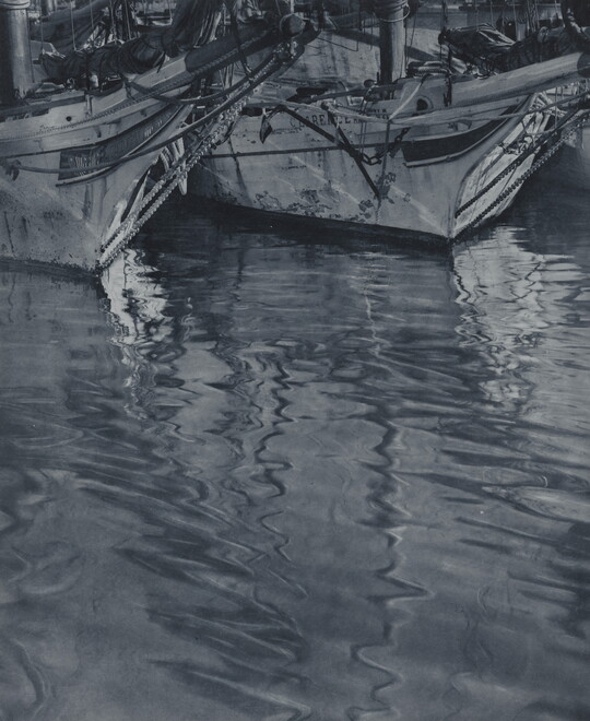 A black-and-white photograph of the ends of two docked sailboats floating on rippling water.