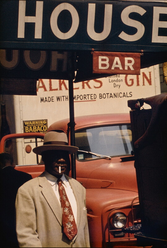 A color photograph of a Black man in a beige hat and suit with a cigarette in his mouth in front of a red car and under a sign that says, "House Bar."