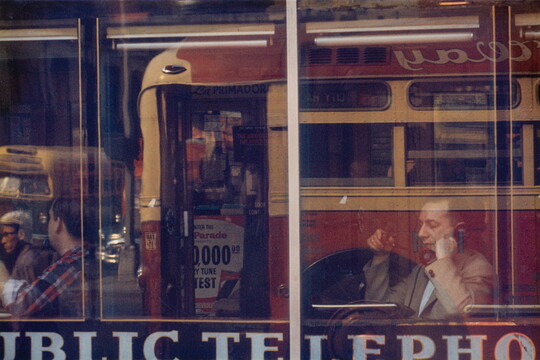 A color photograph of a White man sitting at a cafe window speaking on a phone, with reflections of a passing bus in the glass.