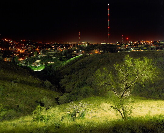 A color photograph of an brightly lit green landscape at night with city lights in the background.
