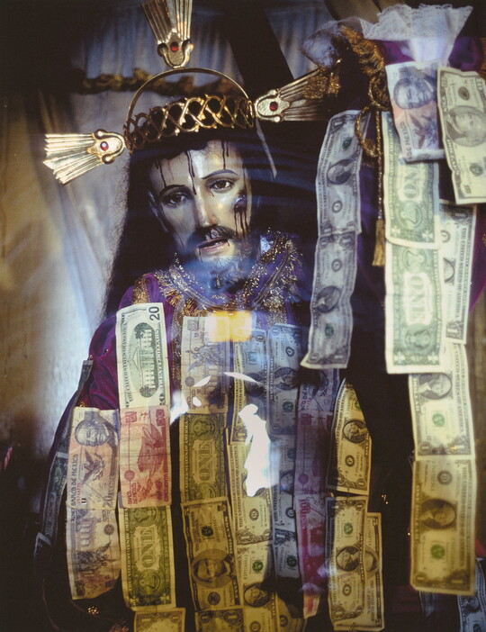 A color photograph of a sculpture of Jesus Christ wearing a crown of thorns behind glass that has strips of dollar bills hanging on it.