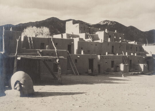 A black-and-white photo of a multi-level adobe pueblo with ladders leading from story to story and dark mountains in the background.
