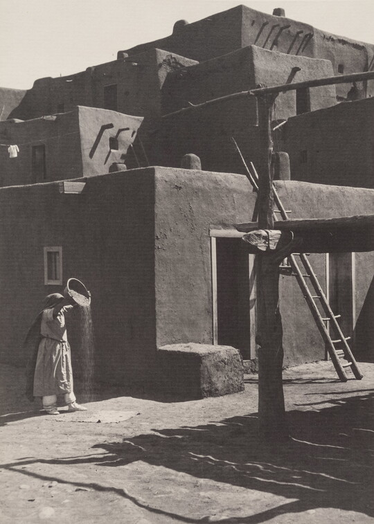 A black-and-white photograph of a woman pouring the contents of a bowl to the ground in front of a multi-level adobe pueblo.