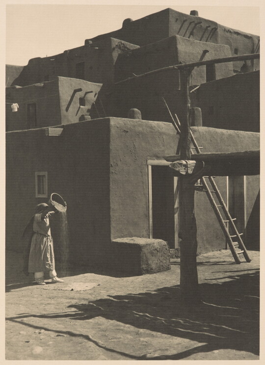 A black-and-white photograph of a woman pouring the contents of a bowl to the ground in front of a multi-level adobe pueblo.