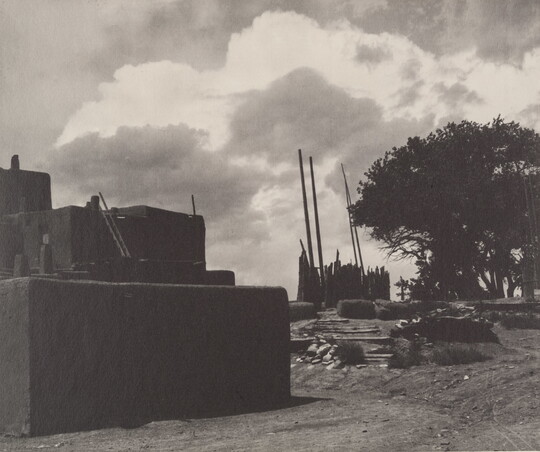 A black-and-white photograph of multi-level adobe pueblo with tall, wood poles standing upright in the distance under a cloudy sky.