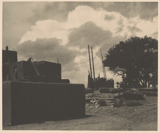 A black-and-white photograph of multi-level adobe pueblo with tall, wood poles standing upright in the distance under a cloudy sky.