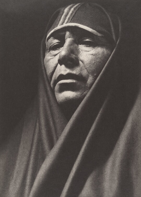 A sepia-toned portrait photograph of an adult Indigenous person with a shawl covering their head and shoulders.