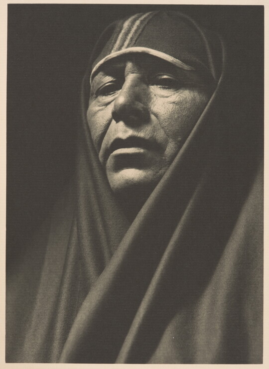 A sepia-toned portrait photograph of an adult Indigenous person with a shawl covering their head and shoulders.