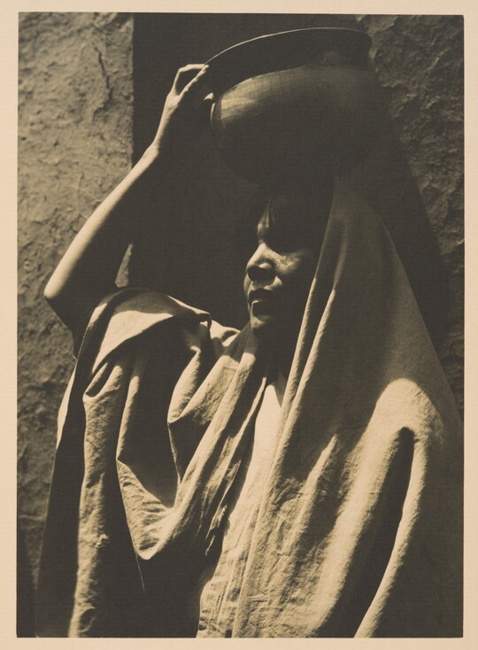 A black-and-white photograph of an Indigenous woman, face mostly in shadow, head and shoulders covered in a blanket or shawl, using her hand to balance a pot on her head.