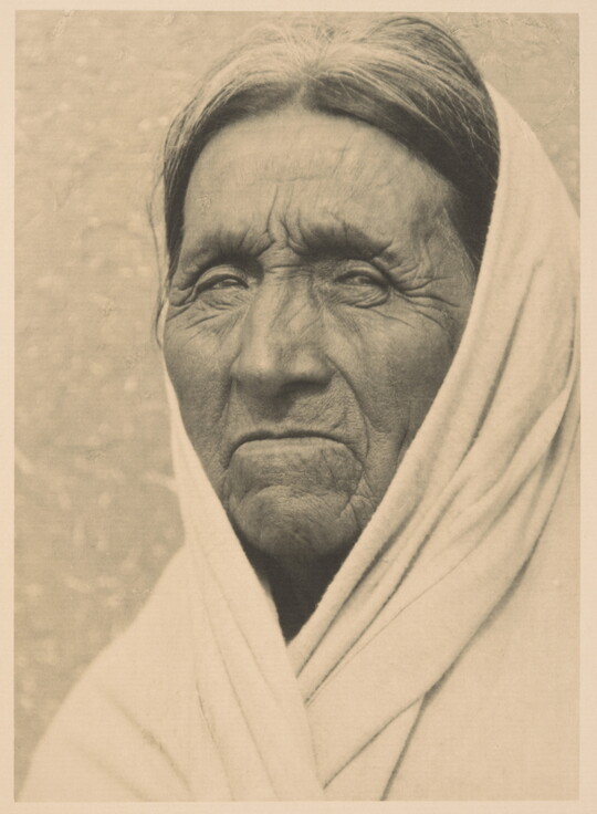 A black-and-white portrait photograph of an elderly Indigenous man with deep wrinkles on his face, a white cloth or shawl covering his head and shoulders.