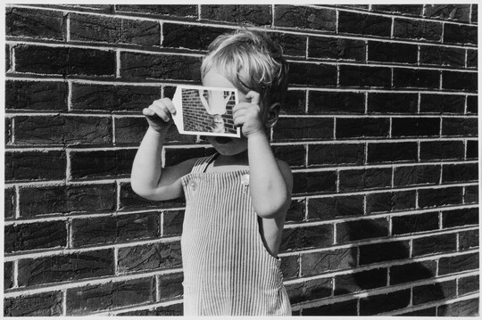 A black-and-white photograph of a White toddler in overalls holding in front of his face an upside down photo of himself standing in front of a brick wall as he stands in front of a brick wall.