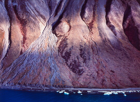 A color photograph of a steep, rocky mountain leading down to a deep blue shore dotted with ice.